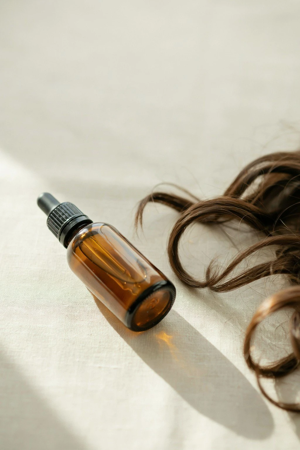 Amber glass dropper bottle of hair oil resting beside loose, healthy-looking hair strands on a light surface