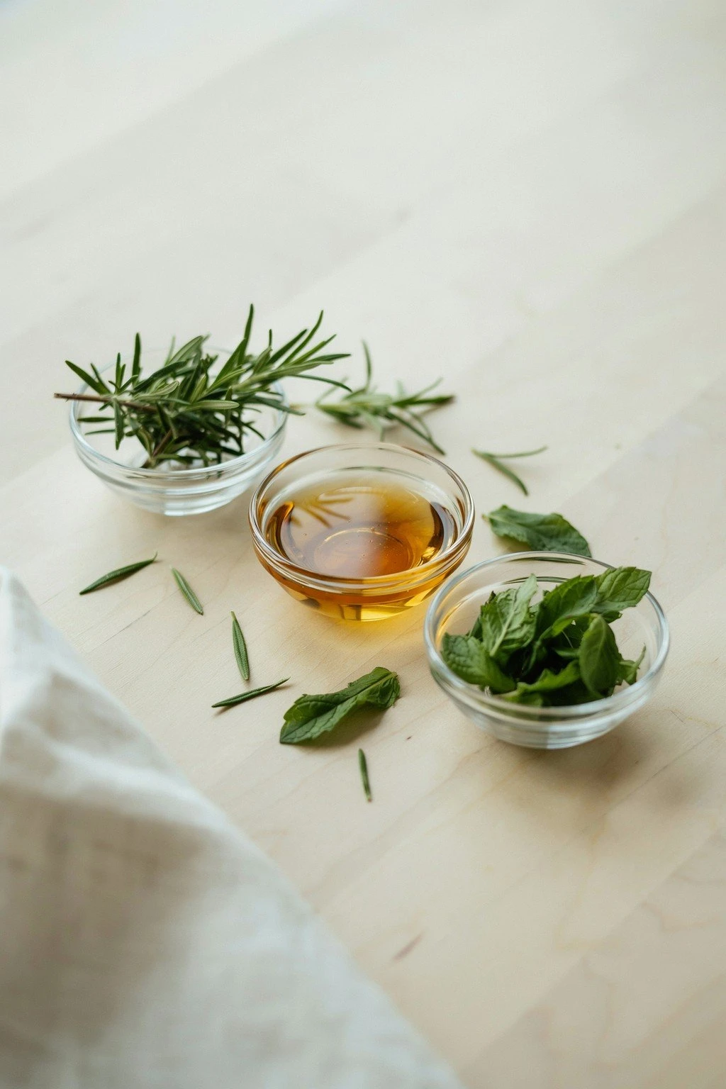 Small glass bowls of castor oil, rosemary sprigs, and peppermint leaves arranged on a wooden surface