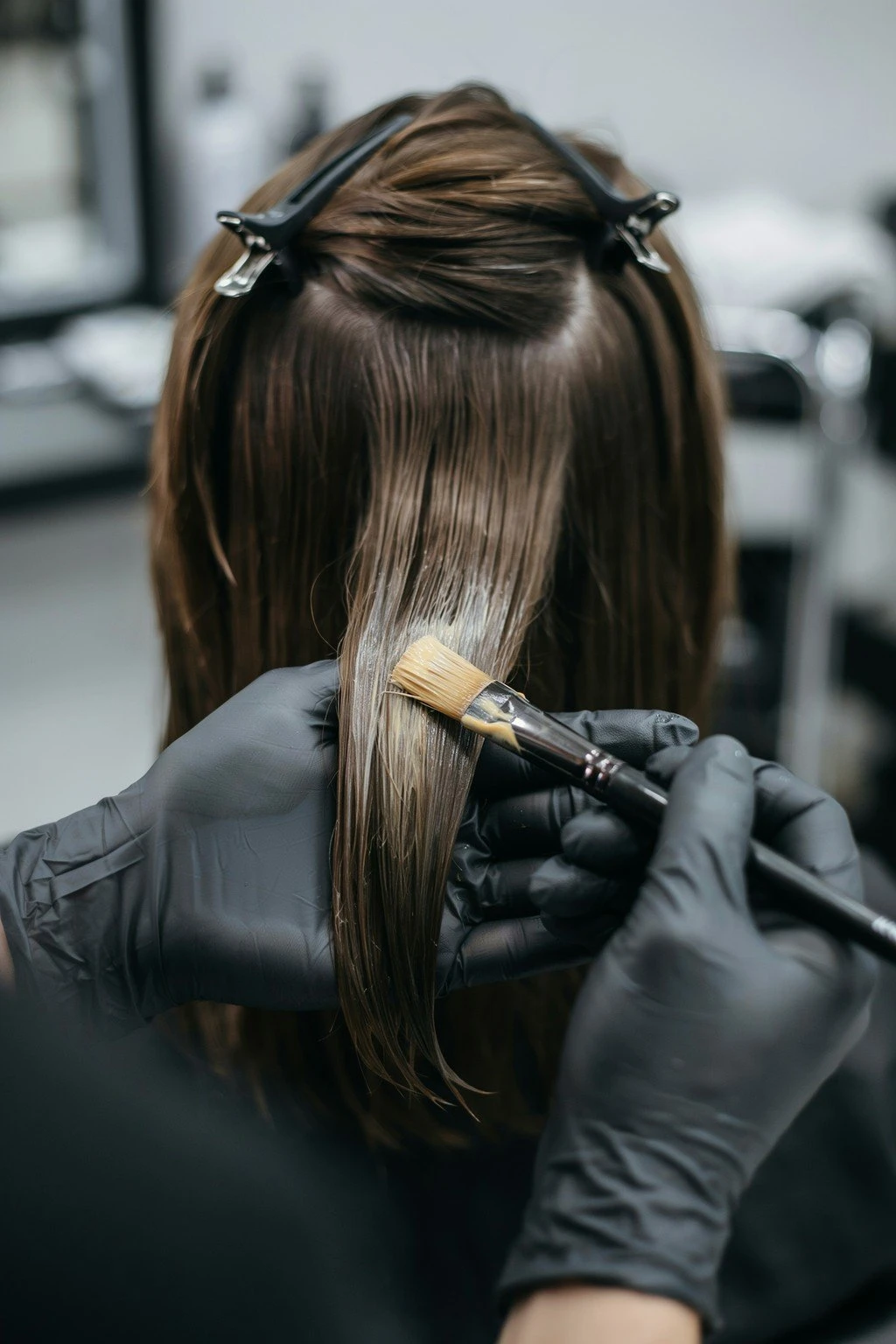 Gloved hands painting lightener freehand onto a section of brunette hair with a flat color brush, no foils visible