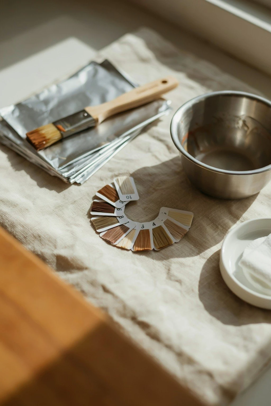 Folded foils, a tint brush, a mixing bowl, and a ring of level swatch cards arranged on a folded salon towel in natural light